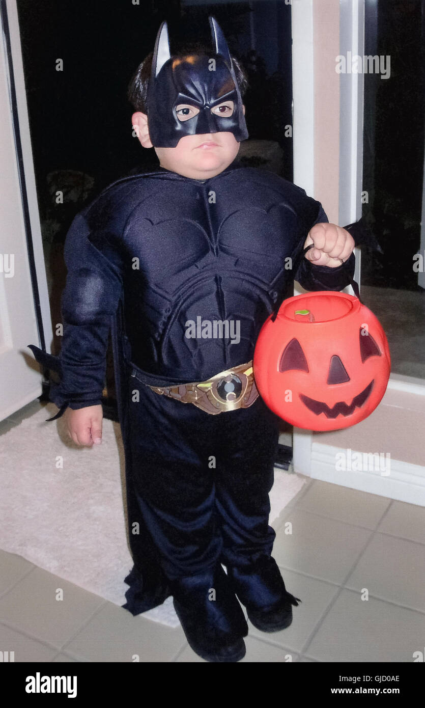 A serious young boy in a Batman costume holds a plastic orange pumpkin  while waiting in a doorway to collect candy from his neighbors on Halloween  eve in California, USA Stock Photo -