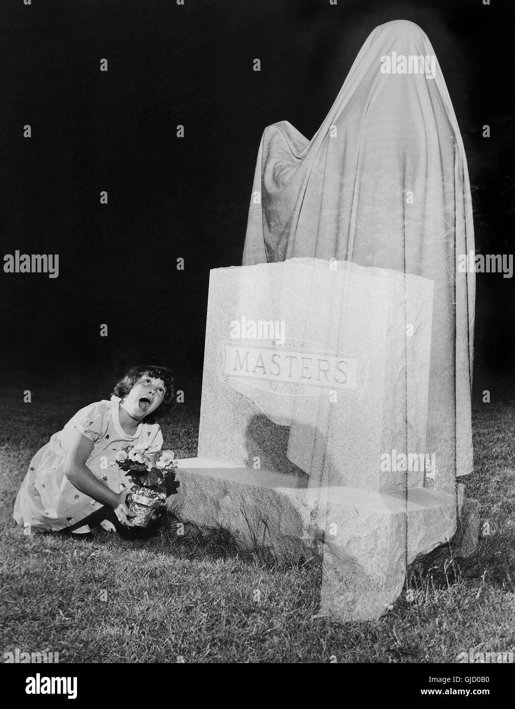 A girl placing flowers at a cemetery grave is surprised by a ghost in this 1960s nighttime photograph created by making a double exposure on the same film frame. The photographer figured a flash exposure setting for the girl and gravestone, and set the lens opening (f/stop) at one-half of that exposure. While the girl held still, a person in a white sheet posed in front of the gravestone and a 2nd flash exposure was made at the same f/stop. Those two half exposures produced a proper exposure. What was recorded during the 1st exposure became visible behind the sheet to create the ghost effect. Stock Photo