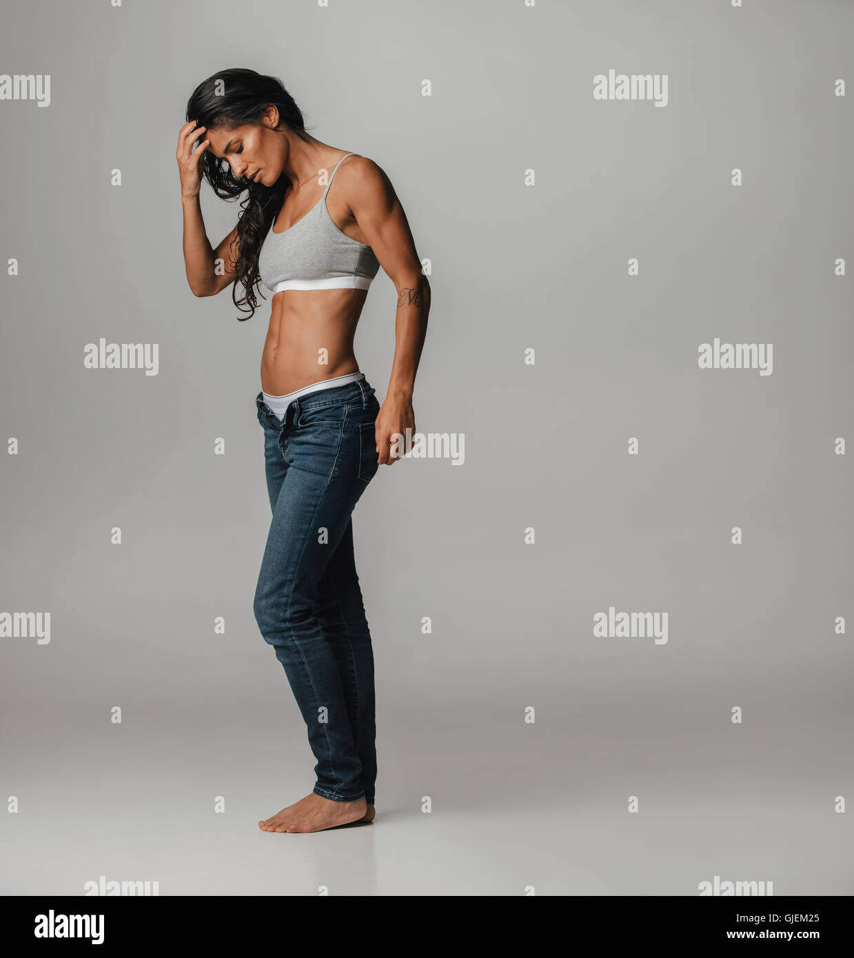 Fit young woman in unbuttoned tight blue jeans and gray sports bra with hand on head looking at feet Stock Photo