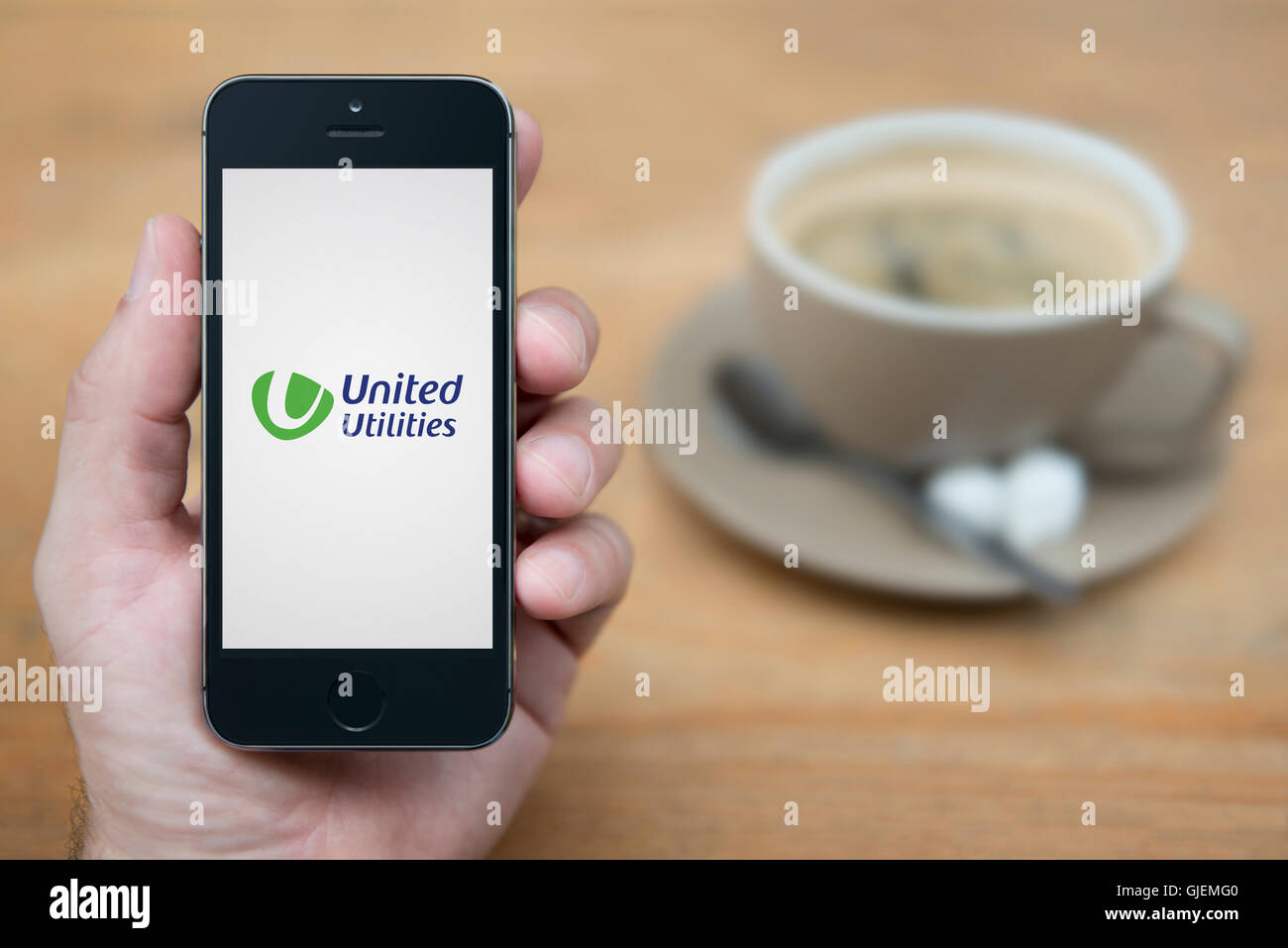 A man looks at his iPhone which displays the United Utilities logo, while sat with a cup of coffee (Editorial use only). Stock Photo