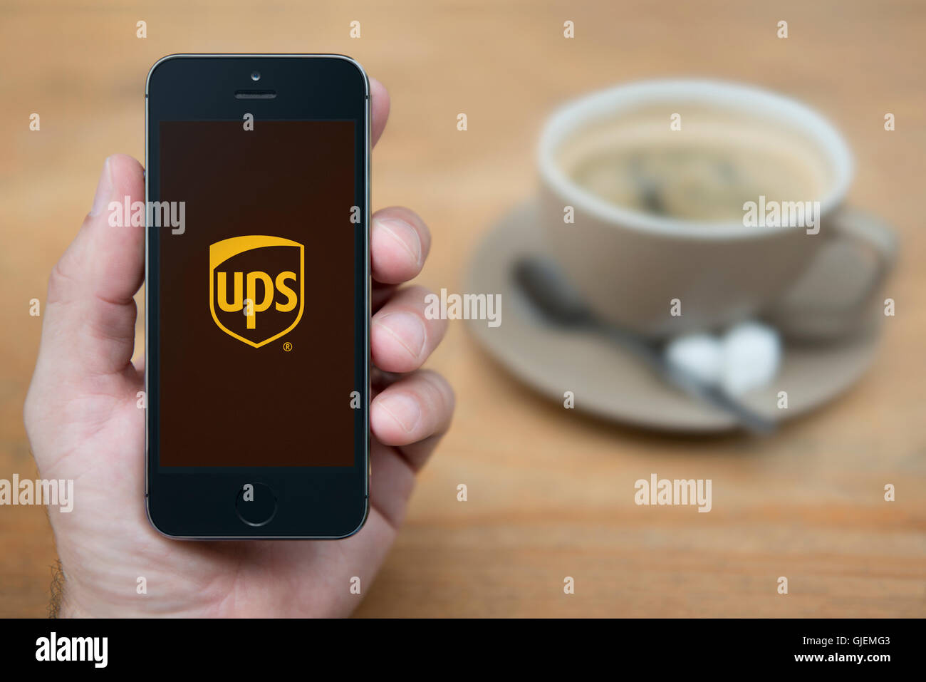 A man looks at his iPhone which displays the UPS logo, while sat with a cup of coffee (Editorial use only). Stock Photo