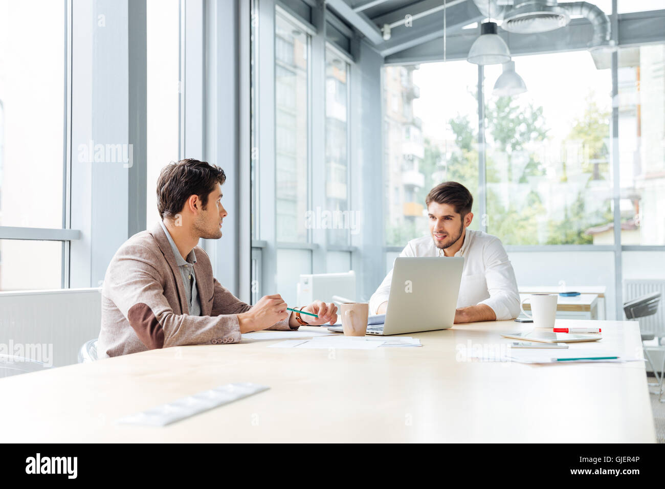Two serious young busnessmen with laptop sitting and talking in office Stock Photo
