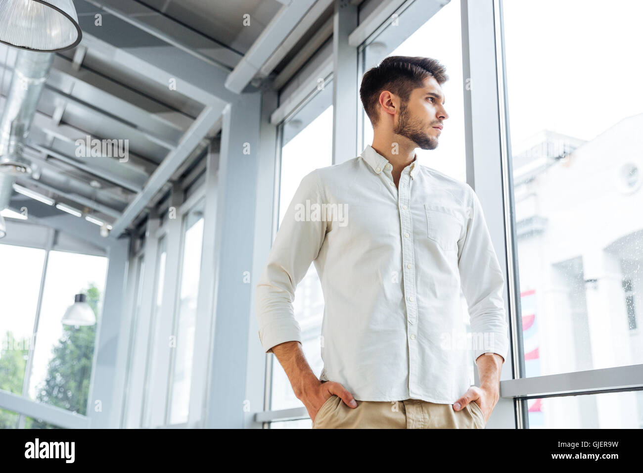 Serious pensive young man standing and thinking near the window Stock Photo