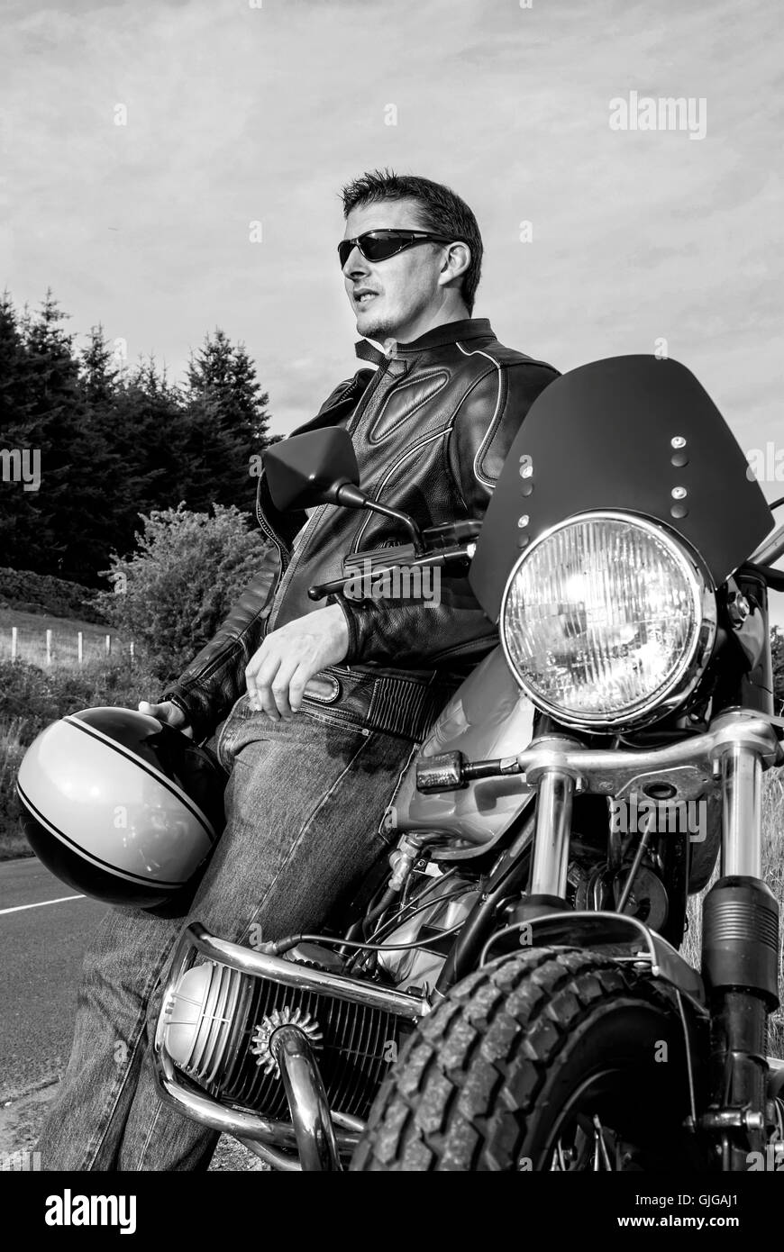 Man motorbiker leaning on motorbike holding helmet leather jacket sunglasses cool guy retro black and white Stock Photo