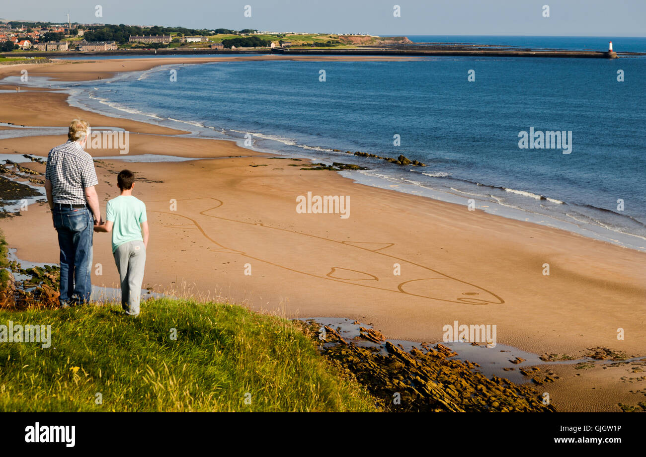 Spittal, Berwick upon Tweed, Northumberland, UK. 16 Aug 2016. Mike Greener and Grandson Lewis Waddell (10) looking down on the biggest salmon the Tweed has ever seen.  Sean Corcoran, 'The Art Hand' from Bunmahon, Co. Waterford, Ireland creates massive sand art drawings with a rake, his first international assignment as a sand artist is Spittal Seaside Festival in Northumberland.  Sean's final piece of work was a Salmon created to remember and celebrate the Salmon Fishers of the River Tweed, his 400 foot long salmon was on the site of Huds Head fishery. Stock Photo
