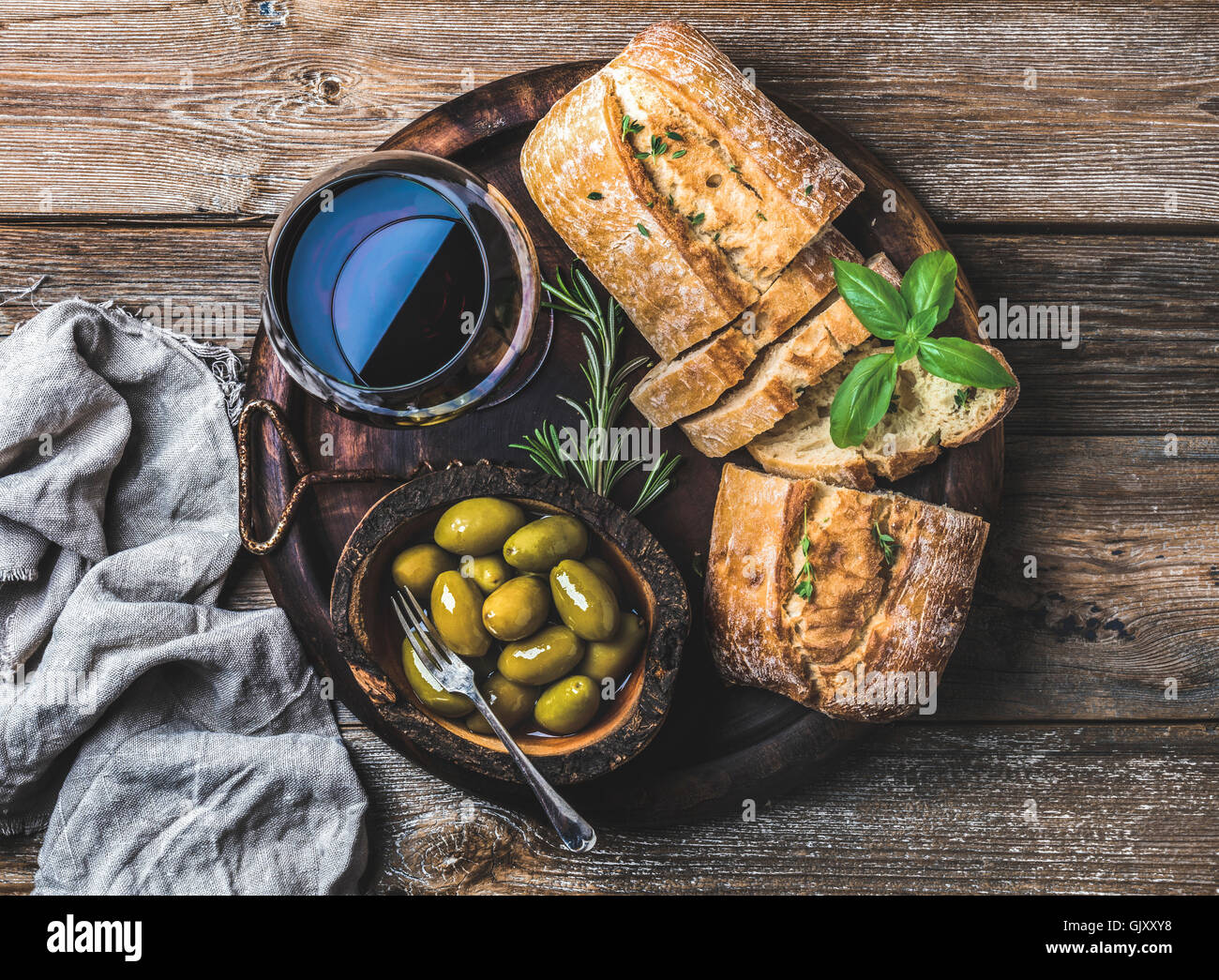 Wine snack set. Glass of red wine, green mediterranean olives, freshly baked ciabatta bread in dark wooden plate over rustic woo Stock Photo