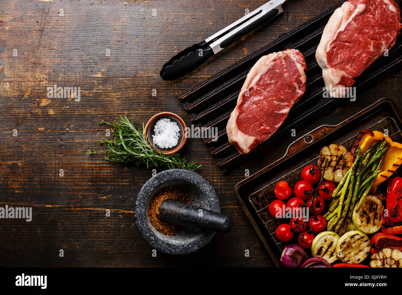 Raw fresh meat Steak Striploin on black cast iron grill and Grilled vegetables on wooden background Stock Photo