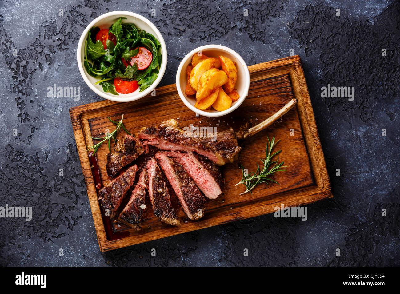 Sliced grilled Medium rare barbecue Steak on bone Veal rib with potato wedges and salad with tomatoes and arugula on dark backgr Stock Photo