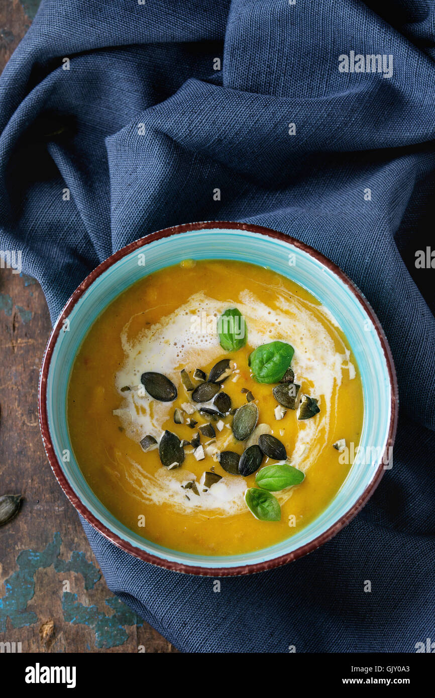 Turquoise ceramic bowl of pumpkin and sweet potato cream soup with fresh basil, cream and seeds, served on textile napkin over o Stock Photo