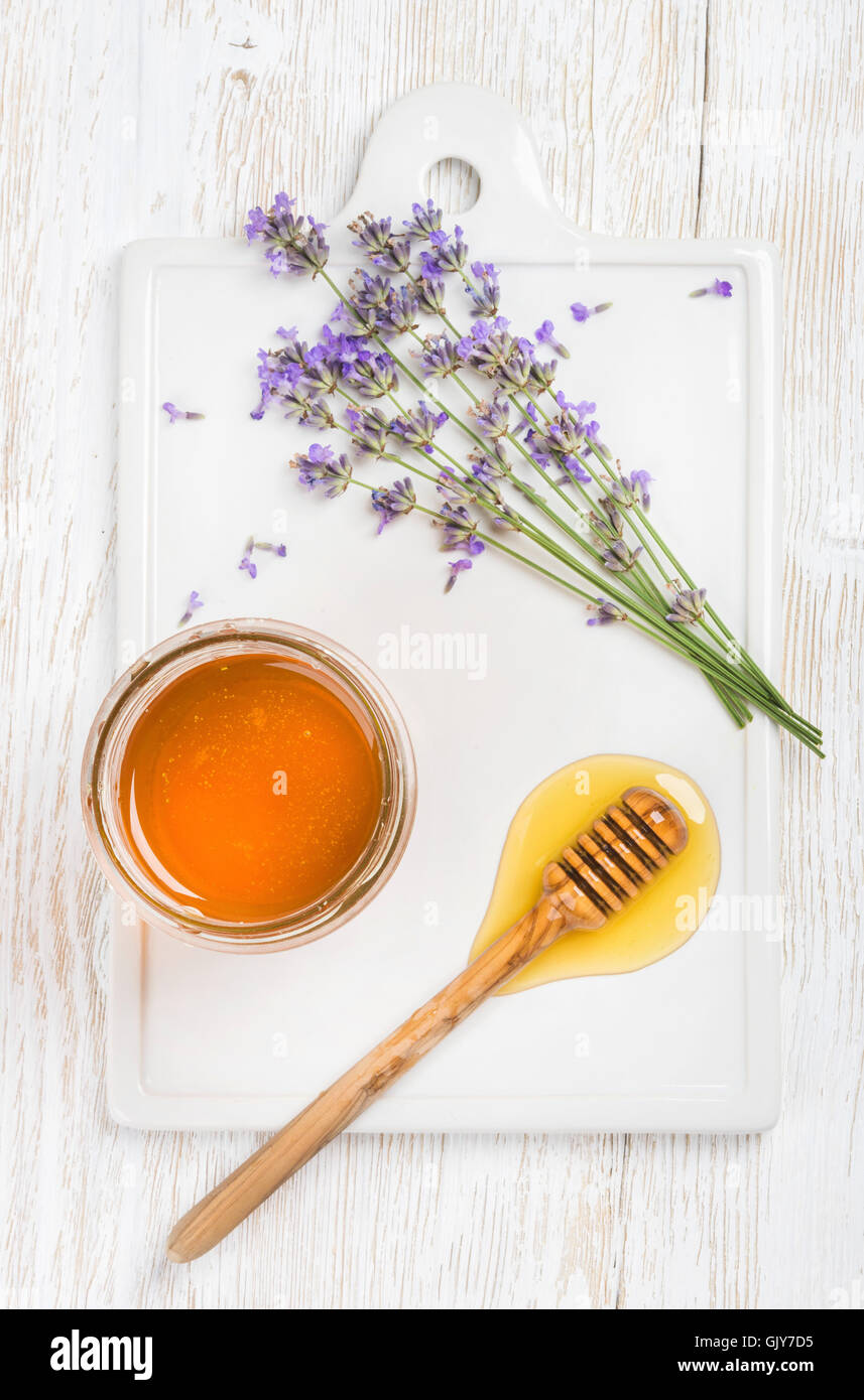 Lavender honey in glass jar served with drizzlier and fresh lavender flowers on ceramic board over white painted wooden backdrop Stock Photo