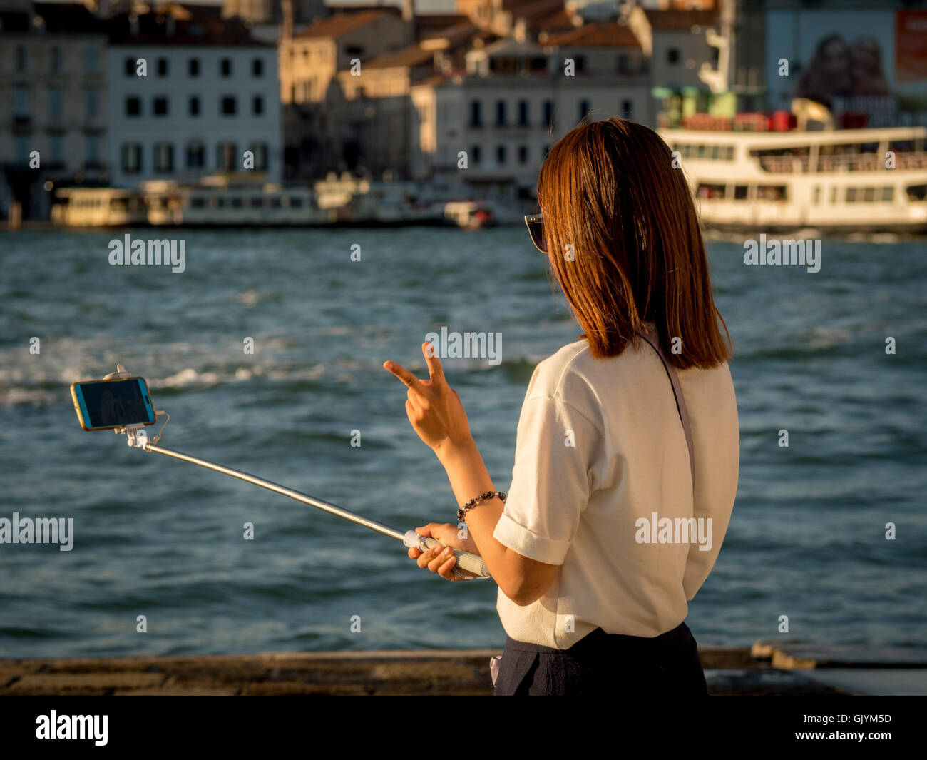 Young Asian adult female tourist taking a selfie, using a selfie stick; making the V sign with her fingers. Venice, Italy. Stock Photo