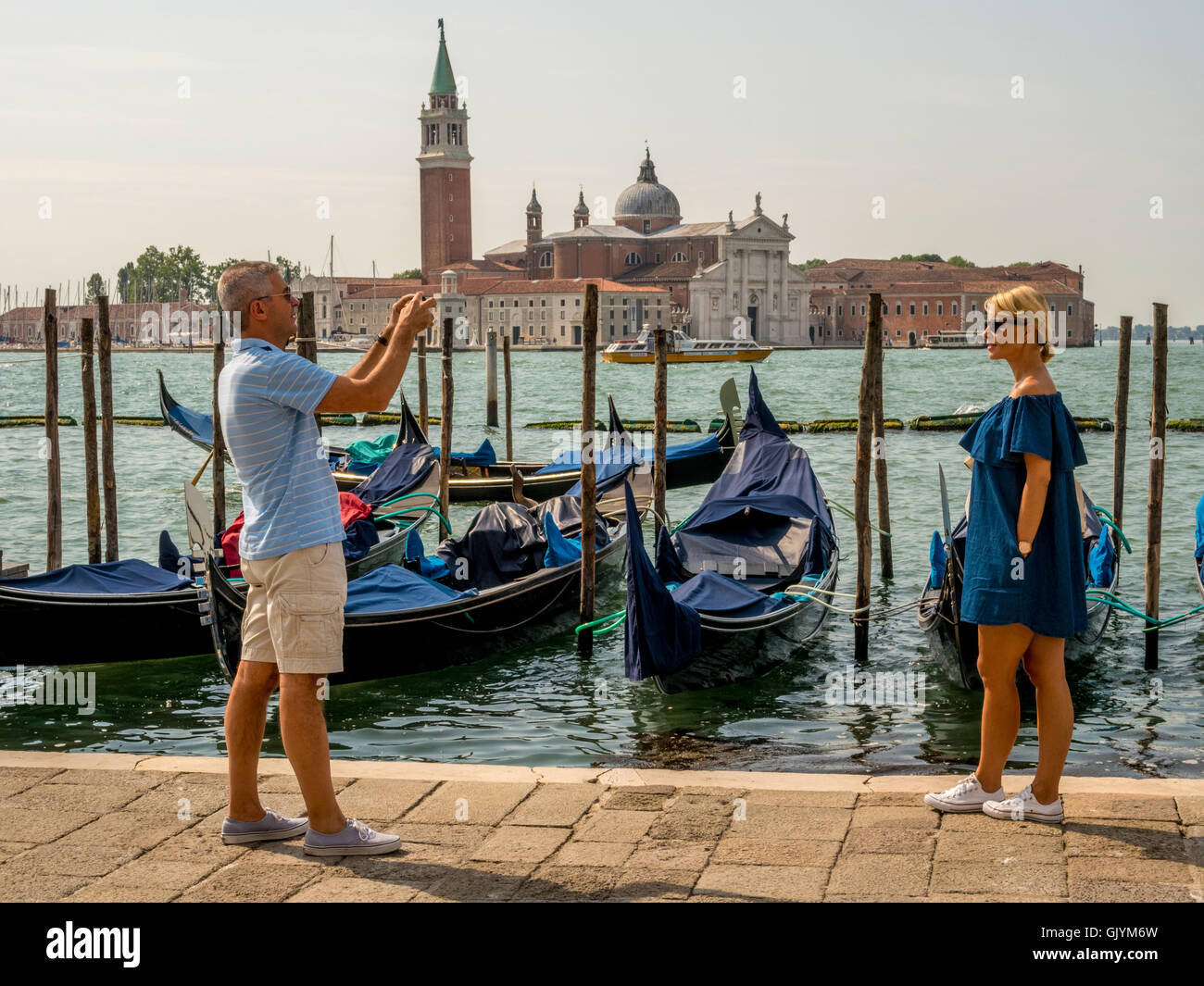 Male adult tourist photographing adult female on the waters edge of Riva degli Schiavoni, with moored gondolas and San Giorgio M Stock Photo