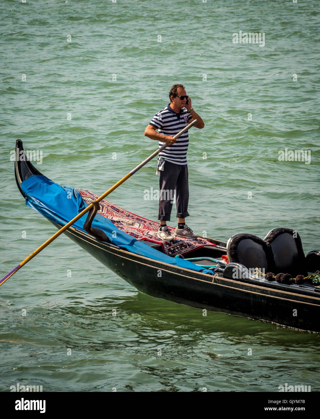 Gondolier wearing traditional striped top steering his gondola whilst talking on his mobile phone. Venice, Italy. Stock Photo