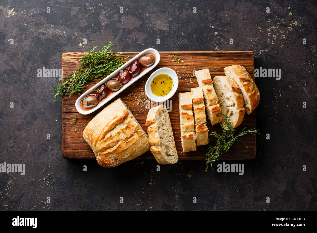 Fresh Ciabatta bread cut in slices on wooden cutting board with olives, oil and herbs on dark background Stock Photo