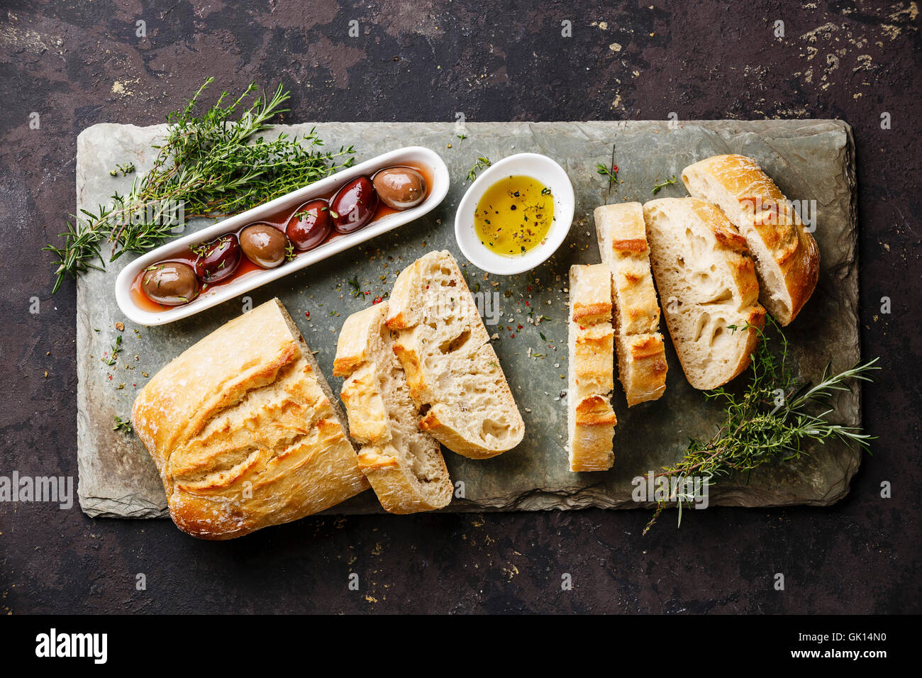 Fresh Ciabatta bread cut in slices on stone slate board with olives, oil and herbs on dark background Stock Photo