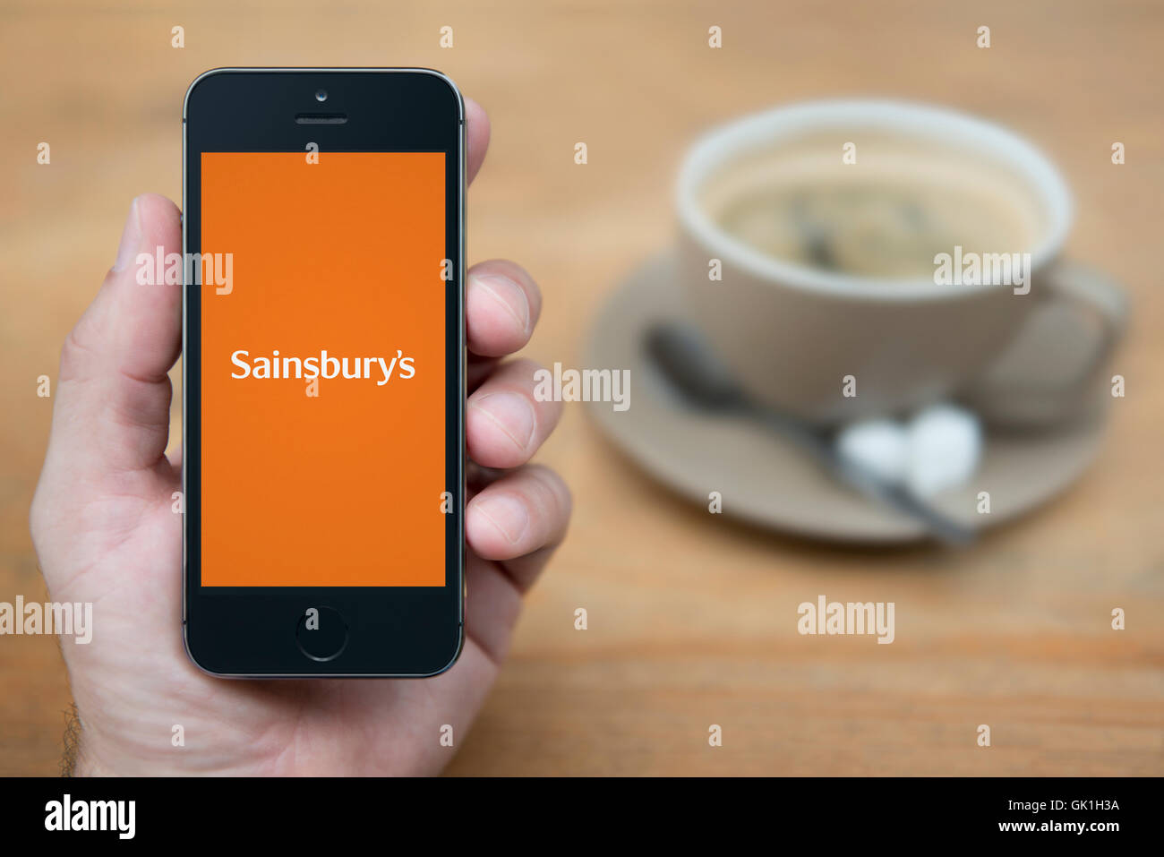 A man looks at his iPhone which displays the Sainsbury's logo, while sat with a cup of coffee (Editorial use only). Stock Photo