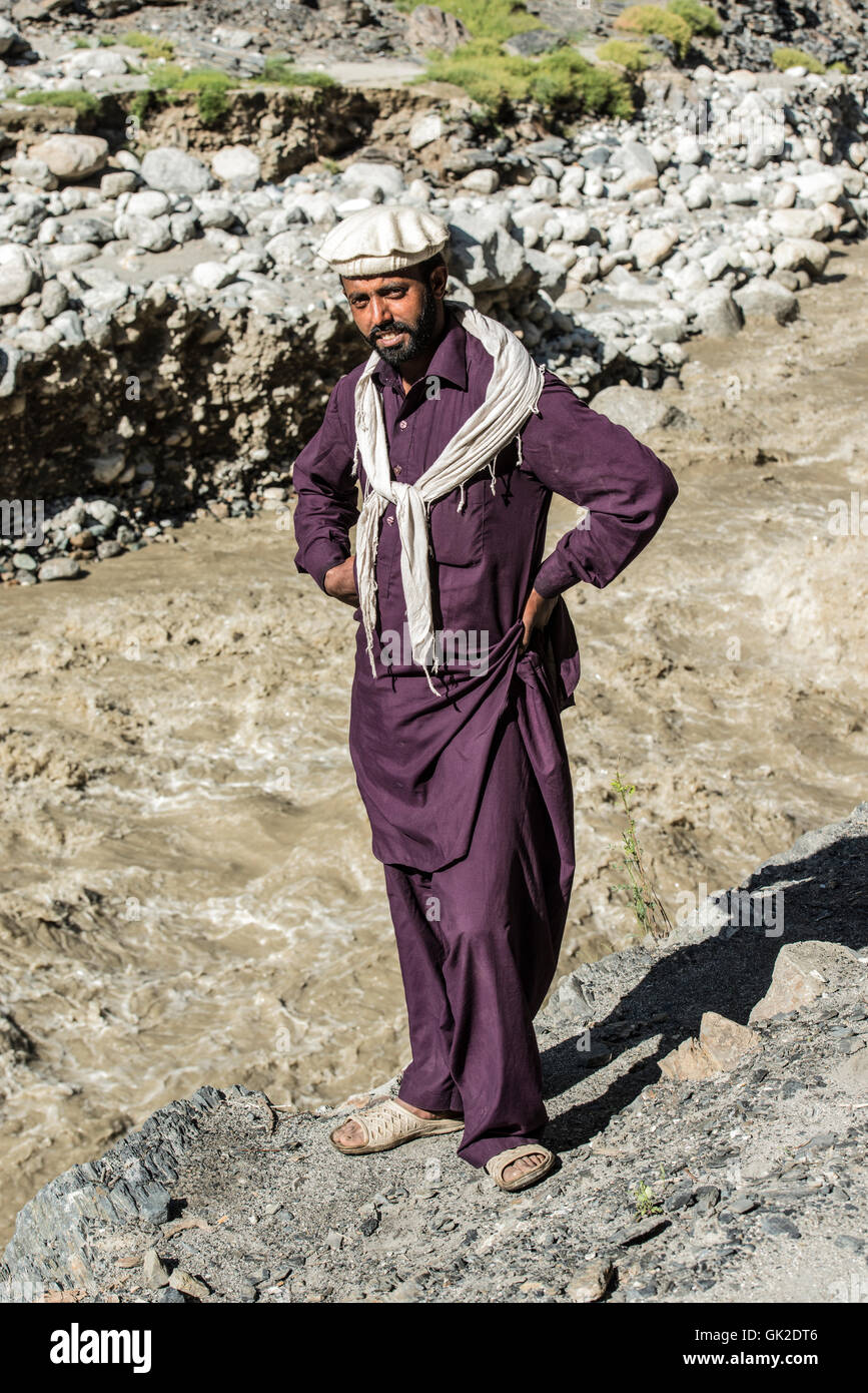 Pakistani man wearing a traditional pagol hat and a purple shalwar kameez standing on the bank of the Kalash river Stock Photo