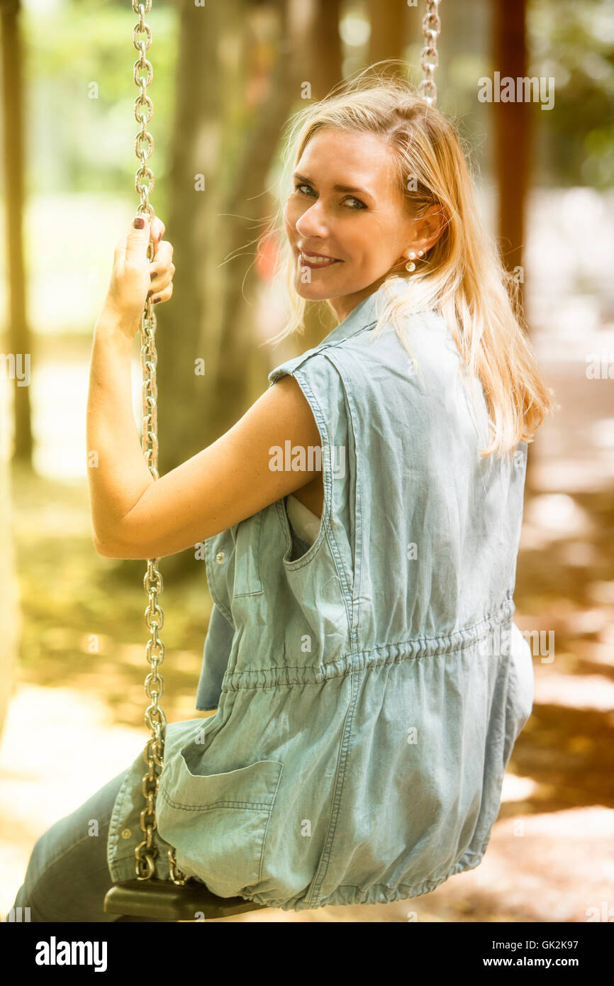 blond woman sitting on a swing at playground and smiles into camera Stock Photo