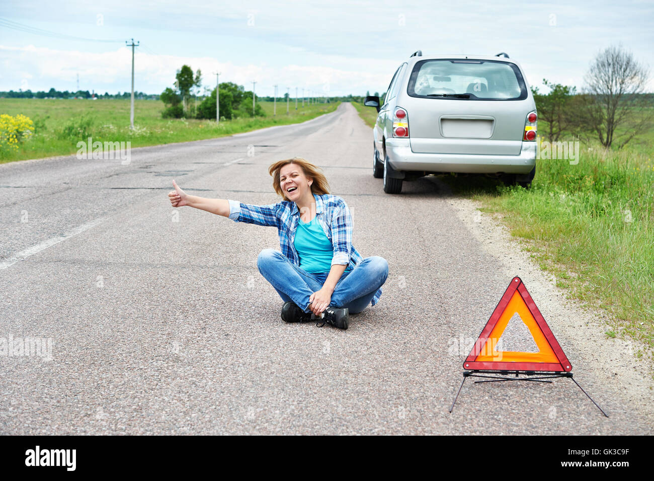 Emotional woman sitting on road near emergency sign and showing thumbs up Stock Photo