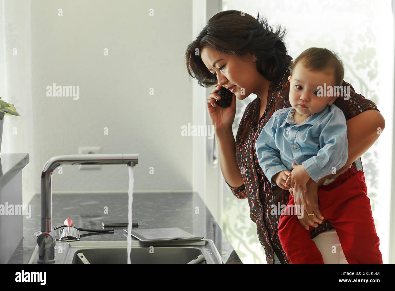 Mother is reading digital tablet while holding her baby on hands and trying to do call and some home work in her kitchen at the Stock Photo