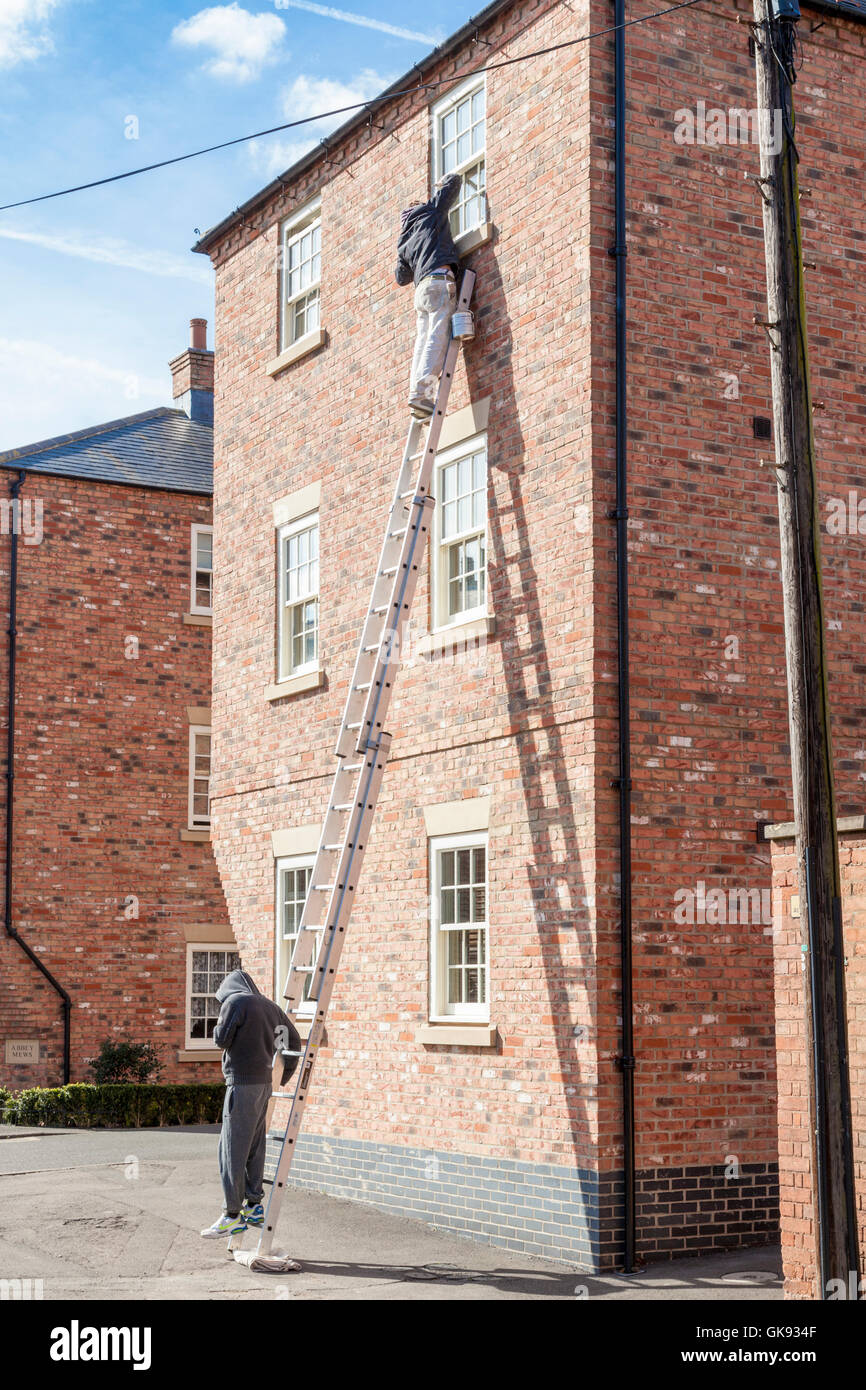 Man on a high ladder painting a house window frame with a person supporting at the bottom, England, UK Stock Photo