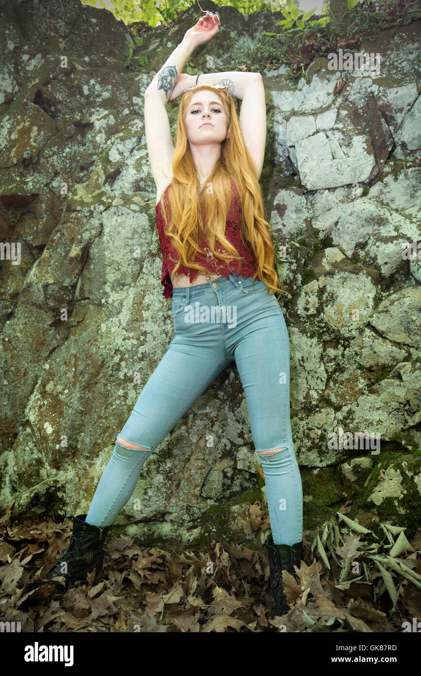 Beautiful red head in skinny jeans, black boots, and red top, leaning full length against traprock cliff with arms raised. Stock Photo