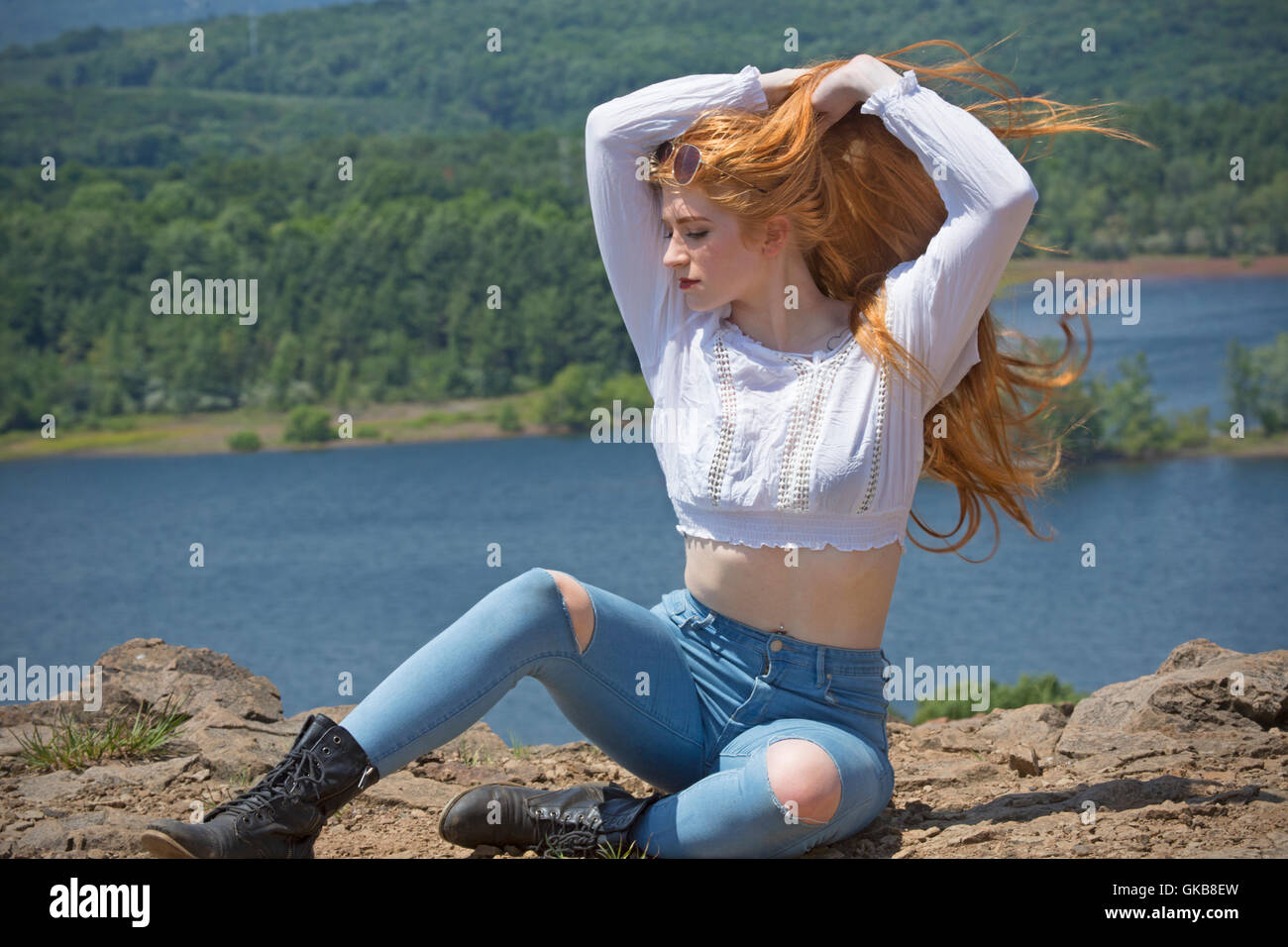 Beautiful red head in skinny jeans, black boots, and white top, sitting on mountain top with wind in hair and a lake below. Stock Photo