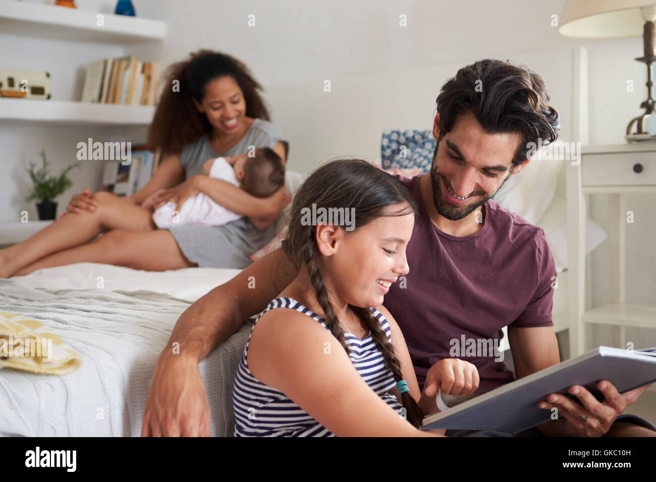 Family With Baby Sitting In Bedroom Reading Book Stock Photo