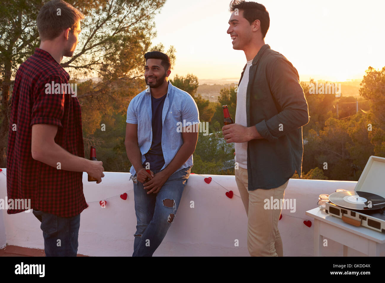 Three male friends talk at a party on a rooftop at sunset Stock Photo