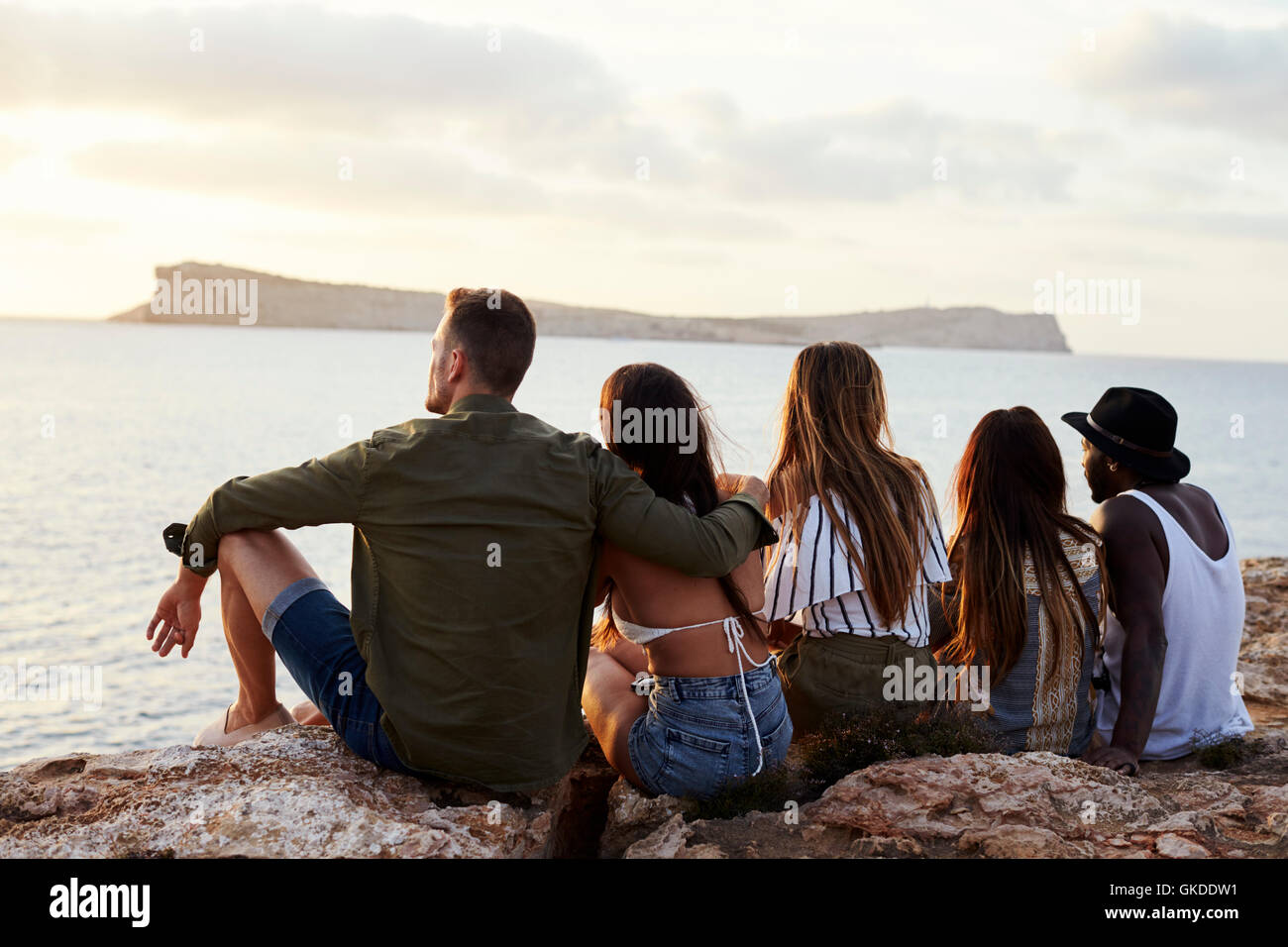 Rear View Of Friends Sitting On Cliff Watching Sunset Stock Photo