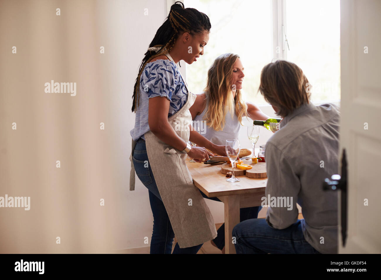 Woman prepares food and friends pour wine, view from doorway Stock Photo