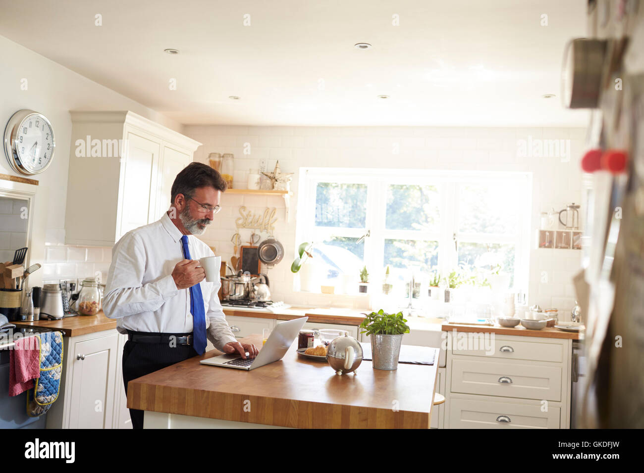 Man in a tie stands holding cup, using laptop in kitchen Stock Photo