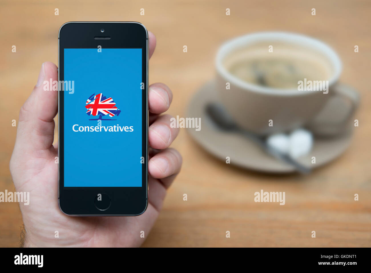 A man looks at his iPhone which displays the Conservatives logo, while sat with a cup of coffee (Editorial use only). Stock Photo