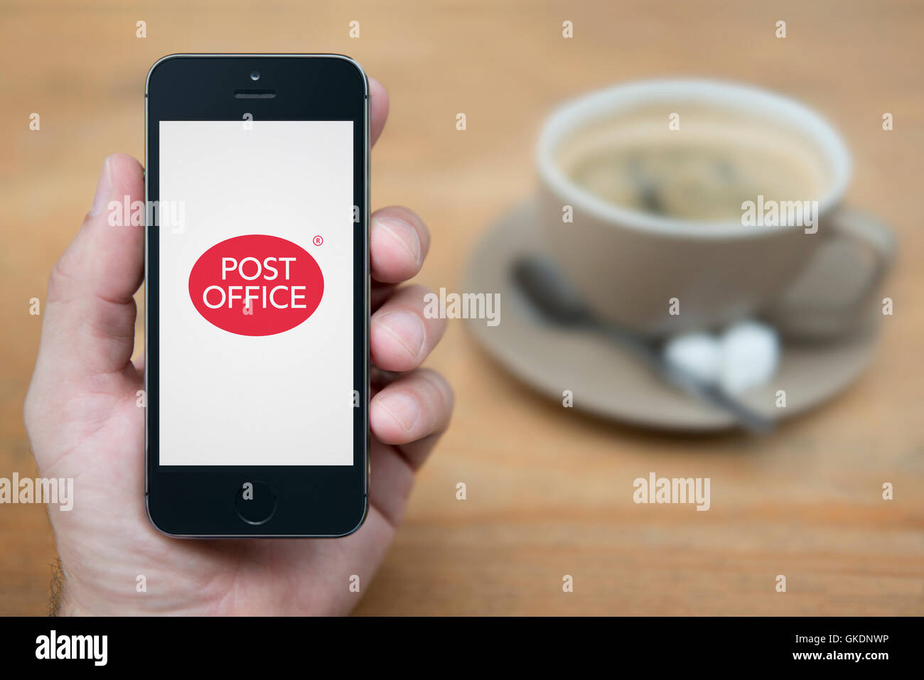A man looks at his iPhone which displays the Post Office logo, while sat with a cup of coffee (Editorial use only). Stock Photo
