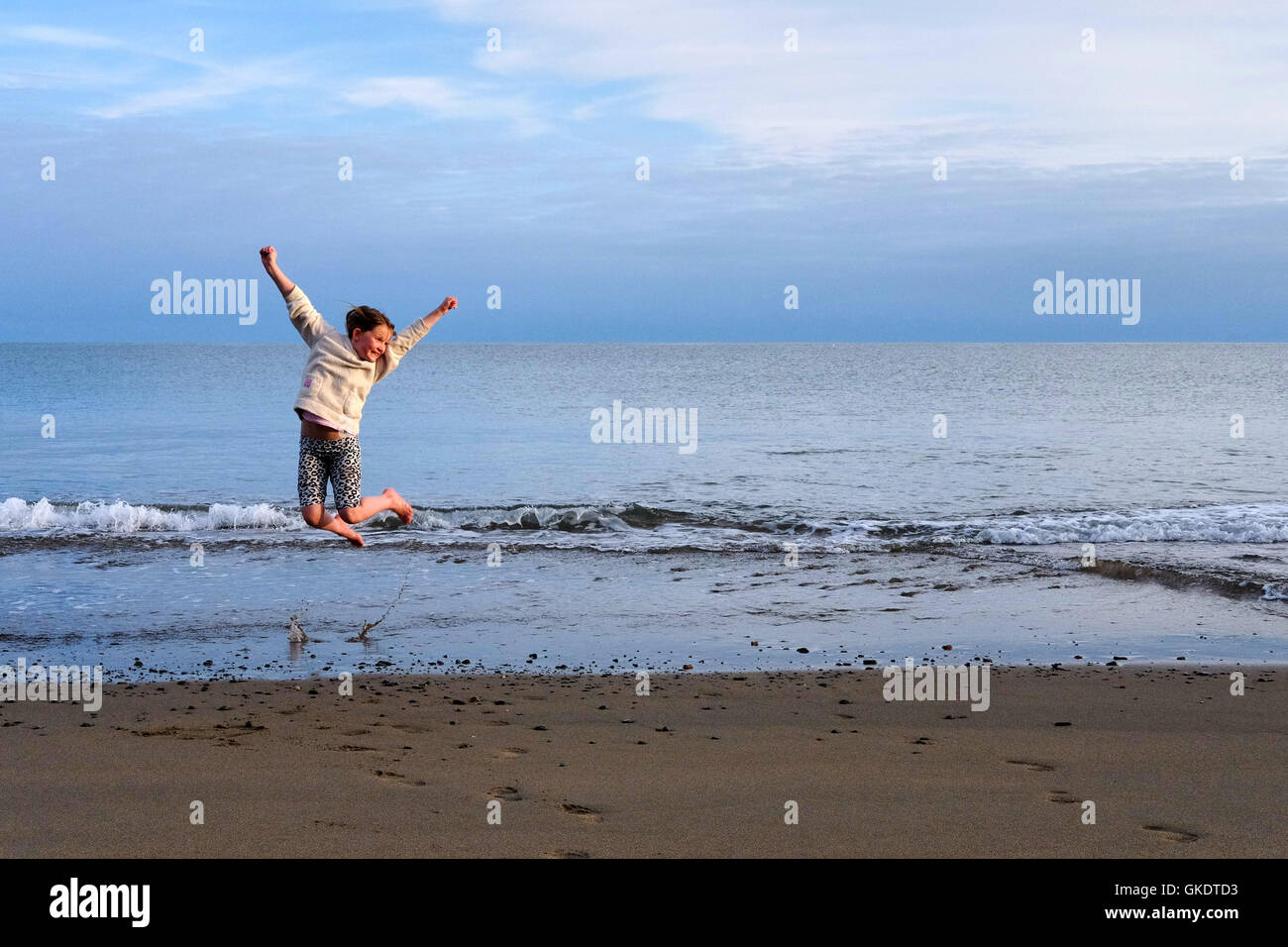 A young girl jumps for joy with arms held high and feet raised on a beach in the evening. Stock Photo