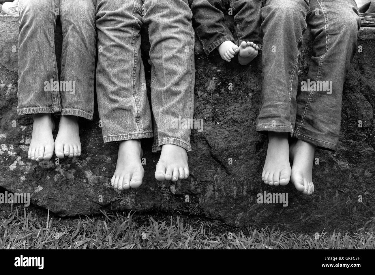 A black and white image of legs and feet of siblings wearing blue jeans and sitting on a rock. Stock Photo