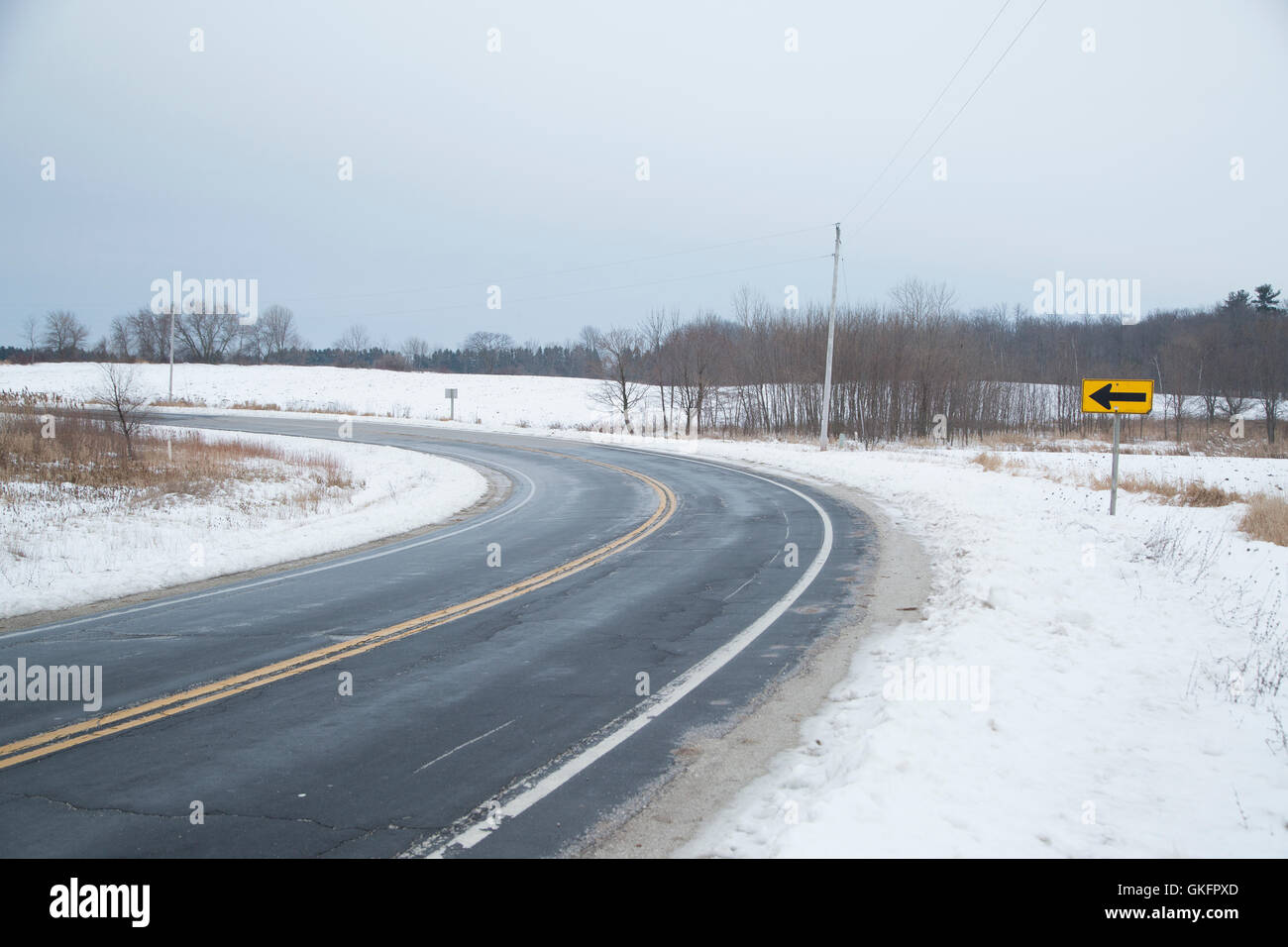 A two lane blacktop road next to a snow covered field curves past an arrow sign on a gray winter day Stock Photo