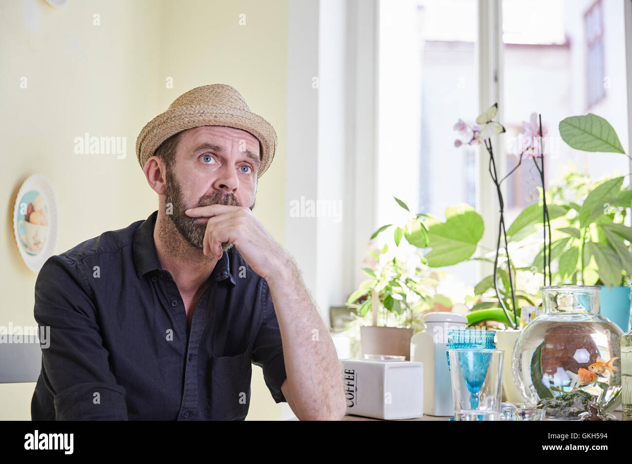 Man with hat and beard sitting and pondering in kitchen Stock Photo