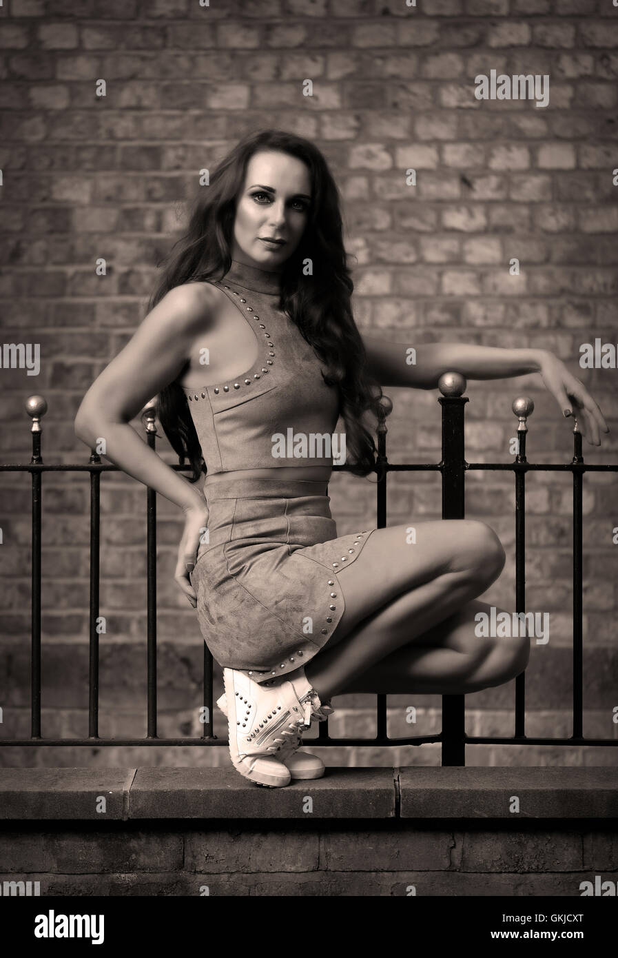 Athletic, muscular female fashion model stands in dance pose against brick wall wearing grey suede stud outfit, white trainers Stock Photo