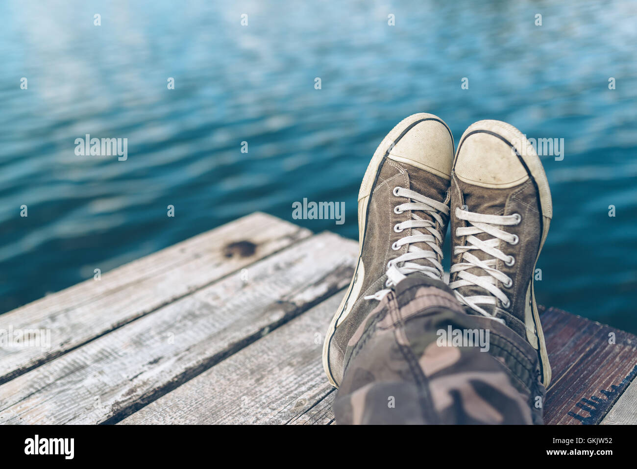 Man with crossed legs relaxing on riverbank pier, casual young guy wearing sneakers sitting by the river in summer afternoon Stock Photo