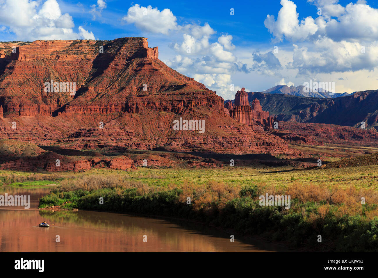 A raft slowly floats around a curve of the Colorado River with an imposing red rock butte and Fisher Towers in the distance Stock Photo