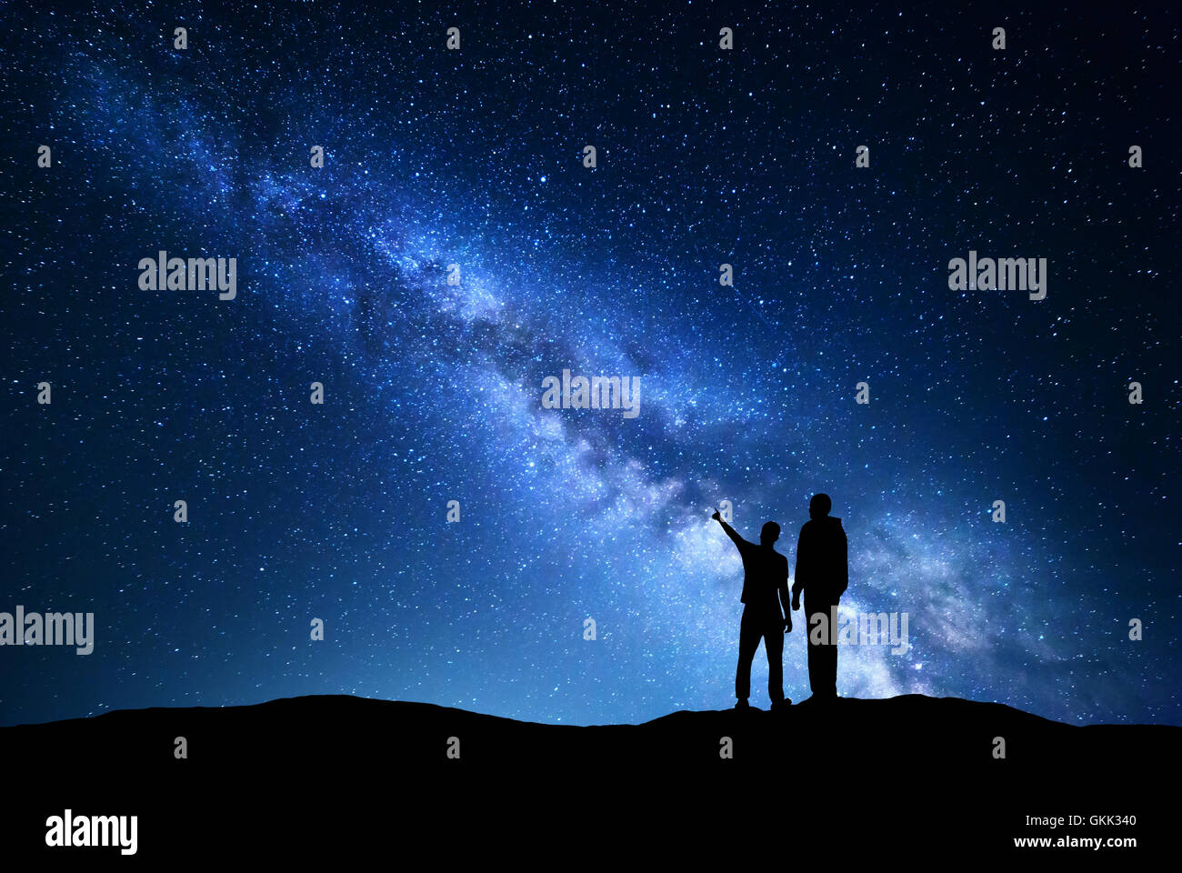 Milky Way with silhouette of men. Father and a son who pointing finger in night starry sky on the mountain. Night landscape. Stock Photo