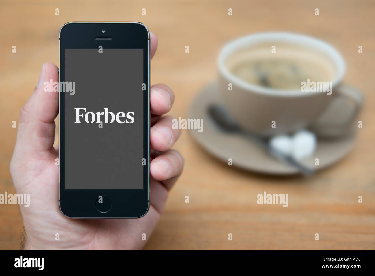 A man looks at his iPhone which displays the Forbes logo, while sat with a cup of coffee (Editorial use only). Stock Photo