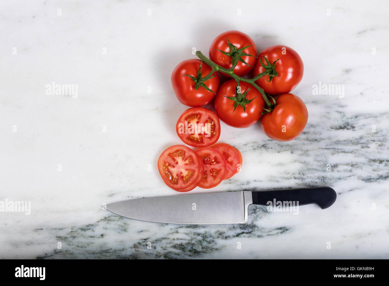 Overhead view of freshly sliced garden tomatoes with large kitchen knife and whole tomatoes on natural marble stone counter. Stock Photo