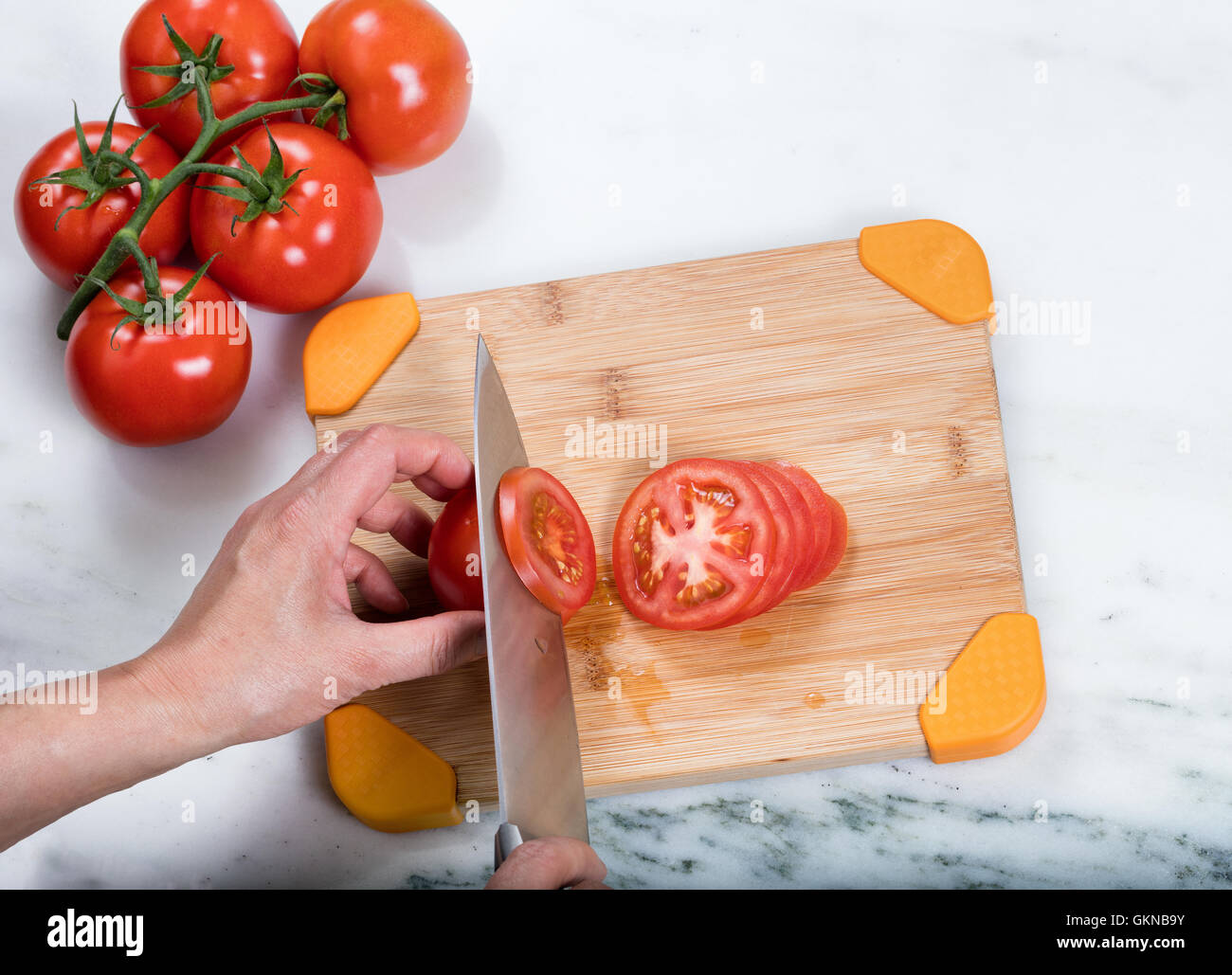 Overhead view of hand slicing a fresh garden tomato with large kitchen knife and whole tomatoes on natural bamboo cutting board. Stock Photo