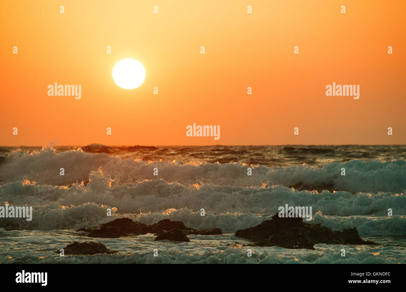 Setting white sun in the orange sky over the ocean with several foaming waves running to the shore Stock Photo