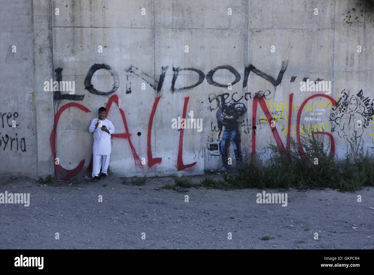 Calais, France. 22nd August 2016. A refugee stands in front of a  graffiti of Apple's Steve Jobs, depicted as a refugee (in allusion to his father, who was a refugee from Syria) that has been painted by Banksy near the entrance of the Jungle.  Over 9,000 refugees live in the Refugee camp in Calais, called the Jungle. They are all on their way to the UK and are stranded here, as the English Channel is heavily guarded with French and English police as well as high fences and barbed wire. Credit:  Michael Debets/Alamy Live News Stock Photo