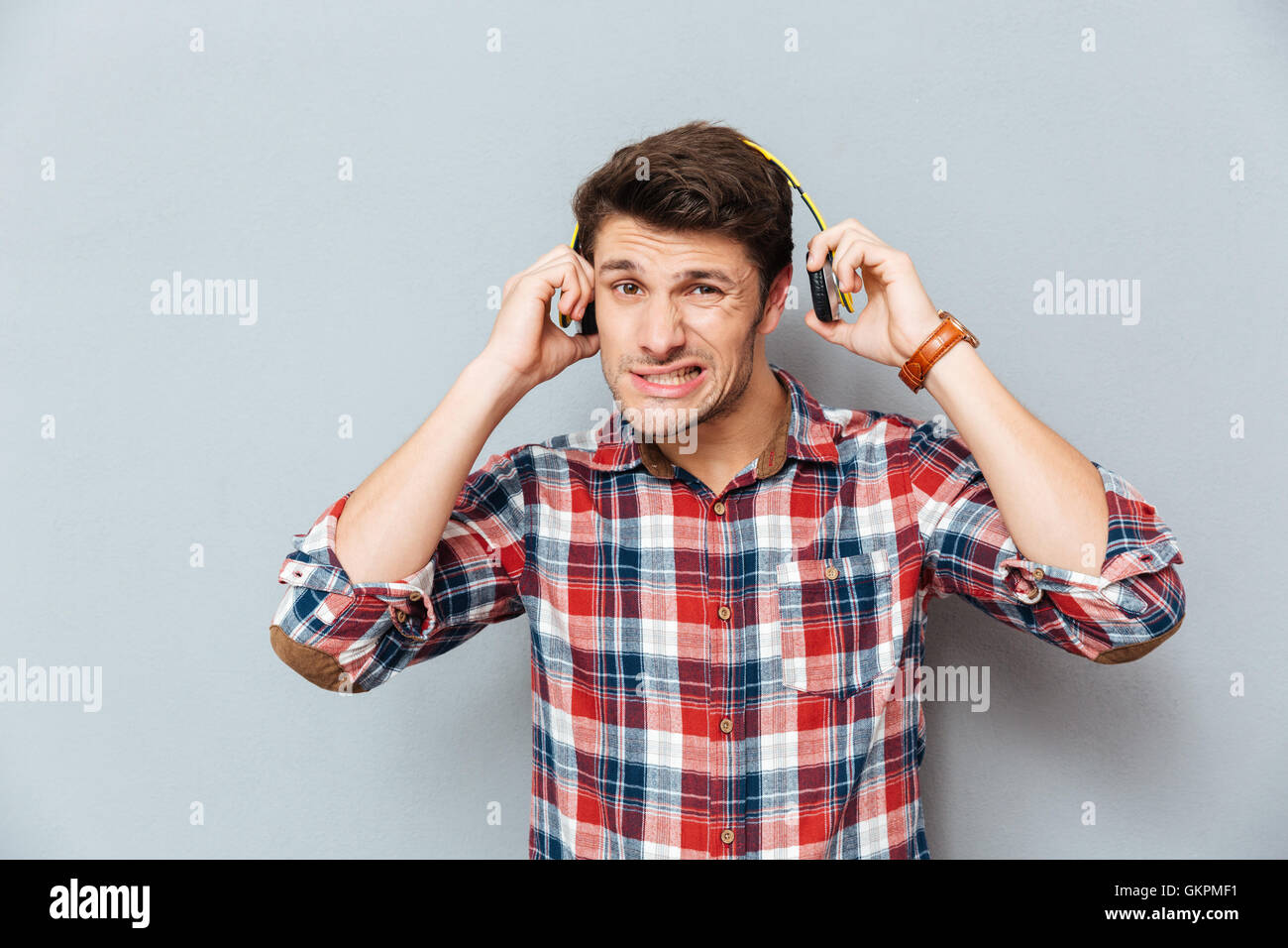 Embarrassed ·irritated young man in checkered shirt taking off headphones over grey background Stock Photo