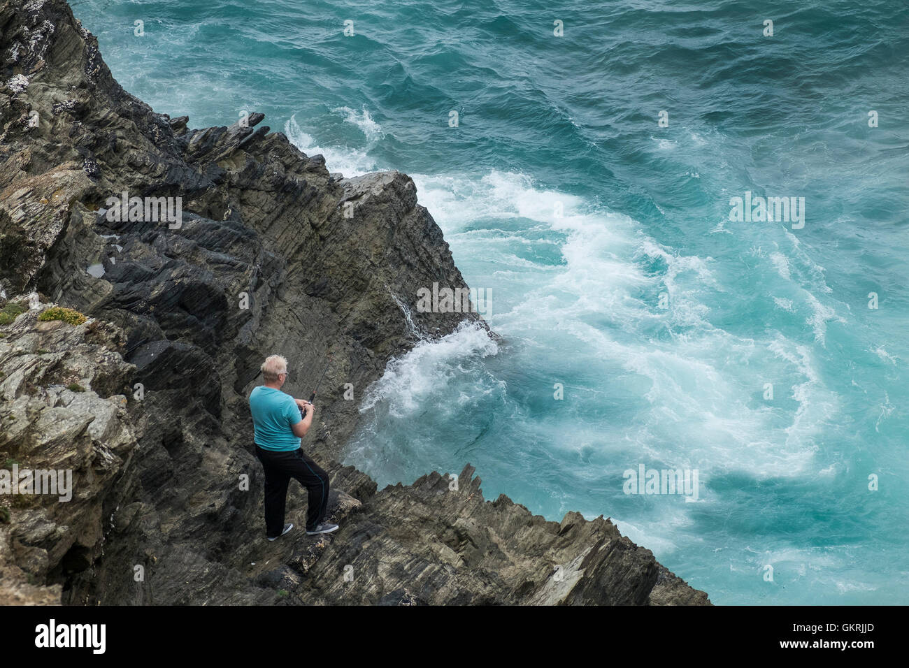 A holidaymaker without any safety equipment fishing from a precarious position on rocks on Towan Headland in Newquay, Cornwall. Stock Photo