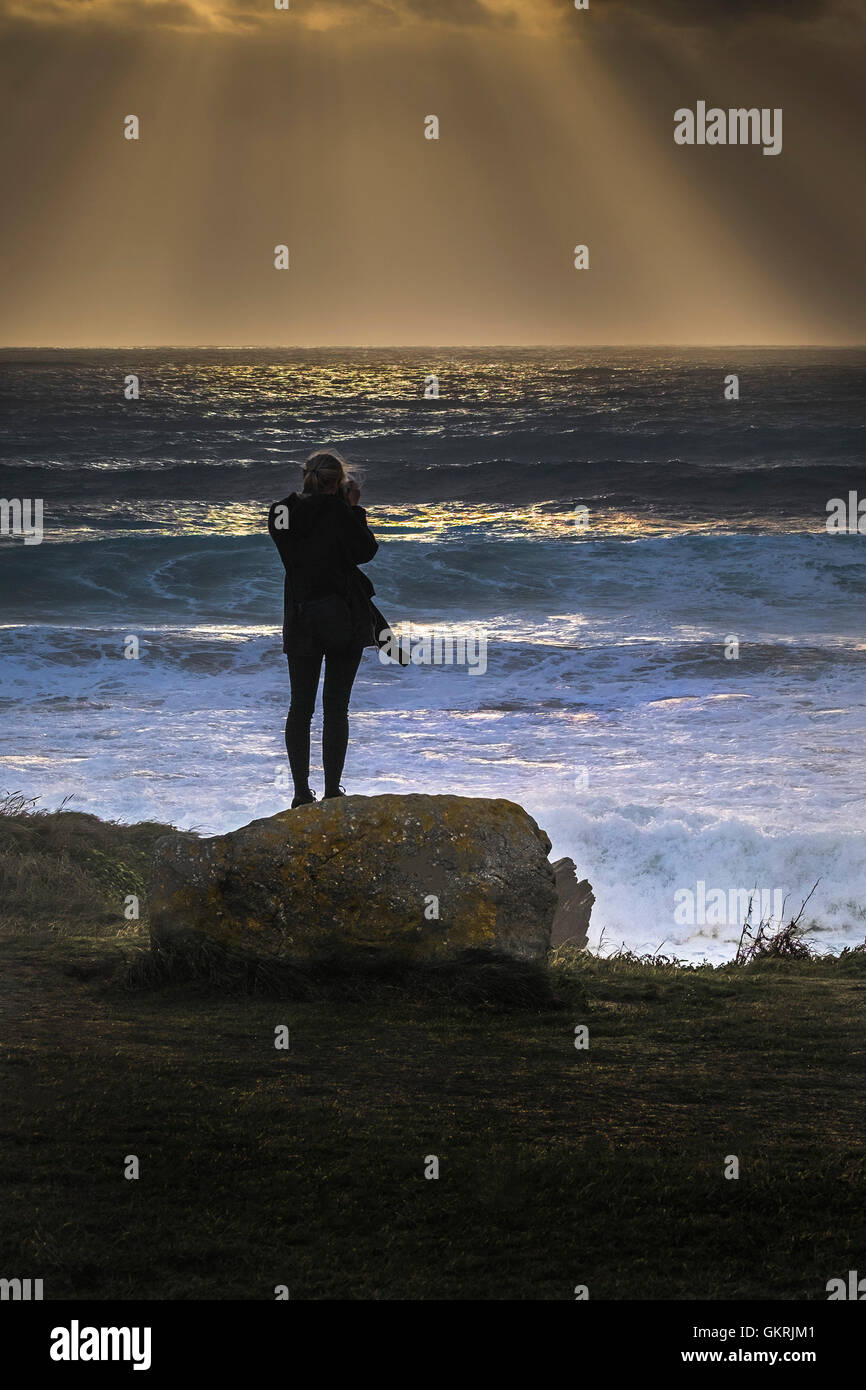A girl stands on a rock and takes a photograph of a spectacular sunset at the Headland in Newquay, Cornwall. Stock Photo